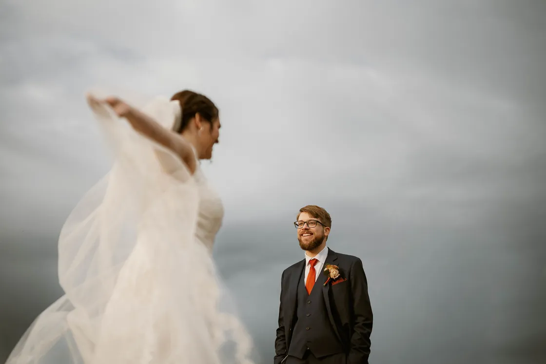 Kelsey's veil catching the wind as Jimmy watches under a gray November sky at their Bishop Farmstead wedding
