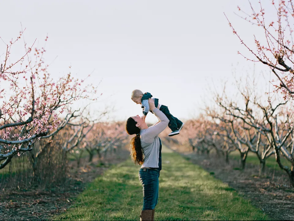 Sandi, studio manager at Twisted Oaks Studio, lifting a laughing toddler in a blossoming peach orchard