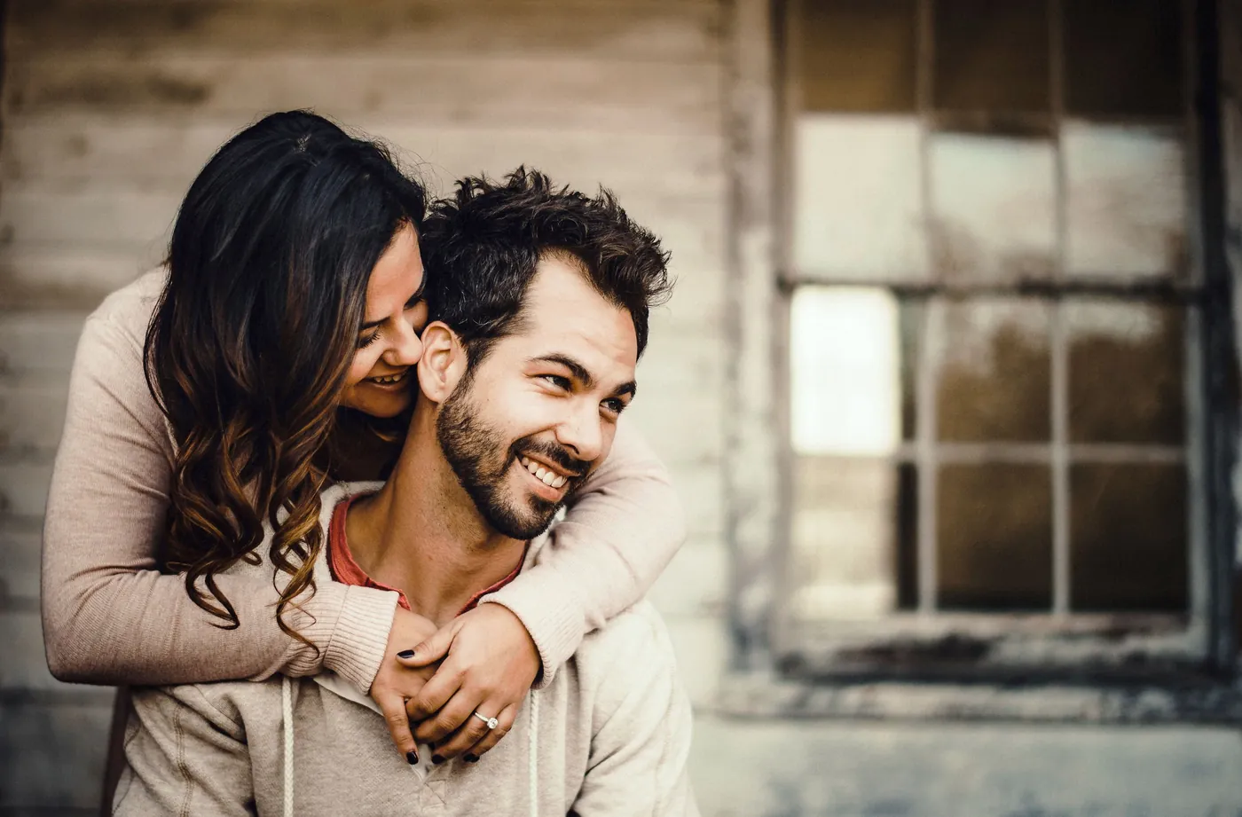Engaged couple laughing as she hugs him from behind in front of a weathered wooden building at Batsto Village in the Pine Barrens, NJ