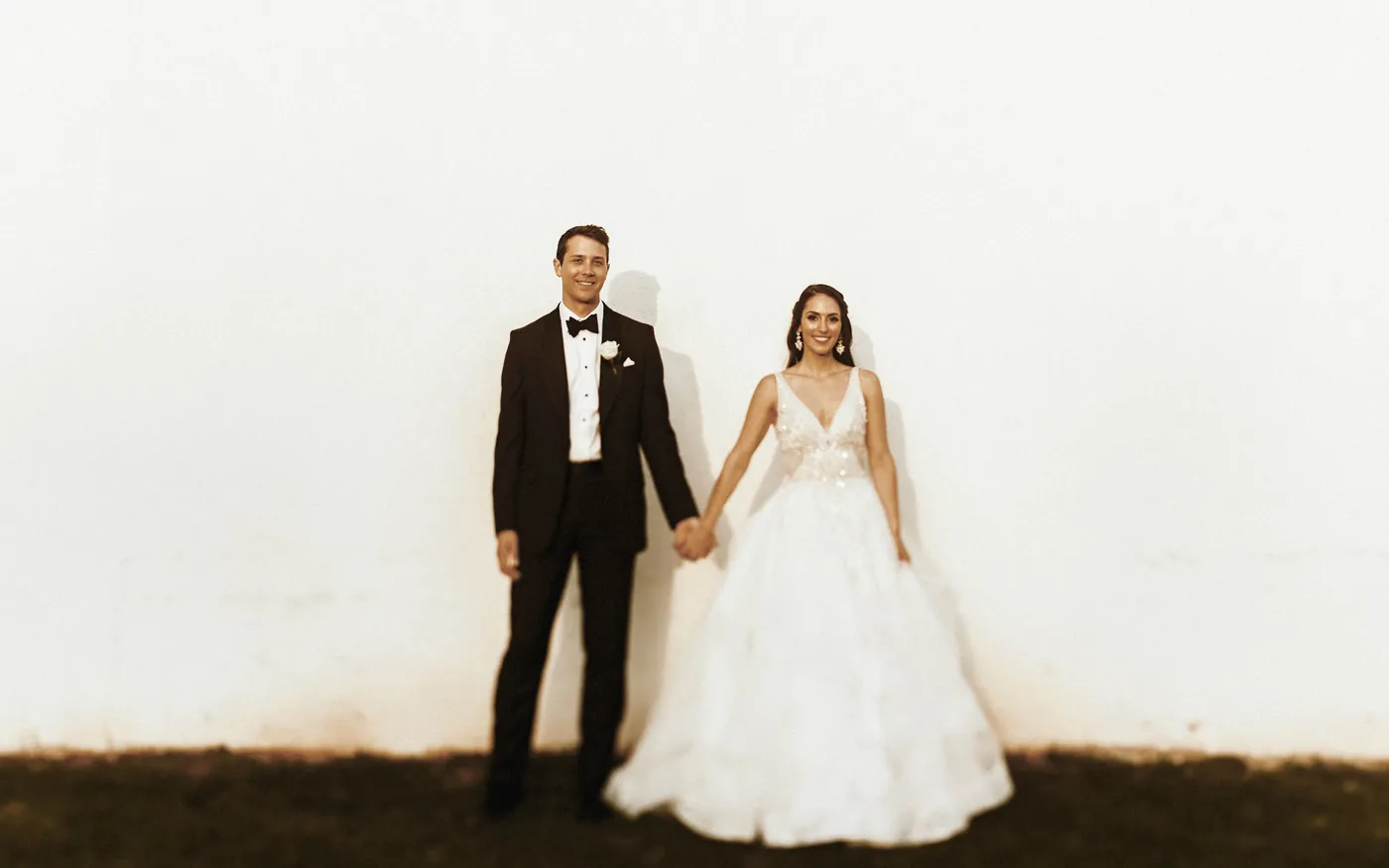 Bride and groom holding hands against a white wall in warm golden hour light