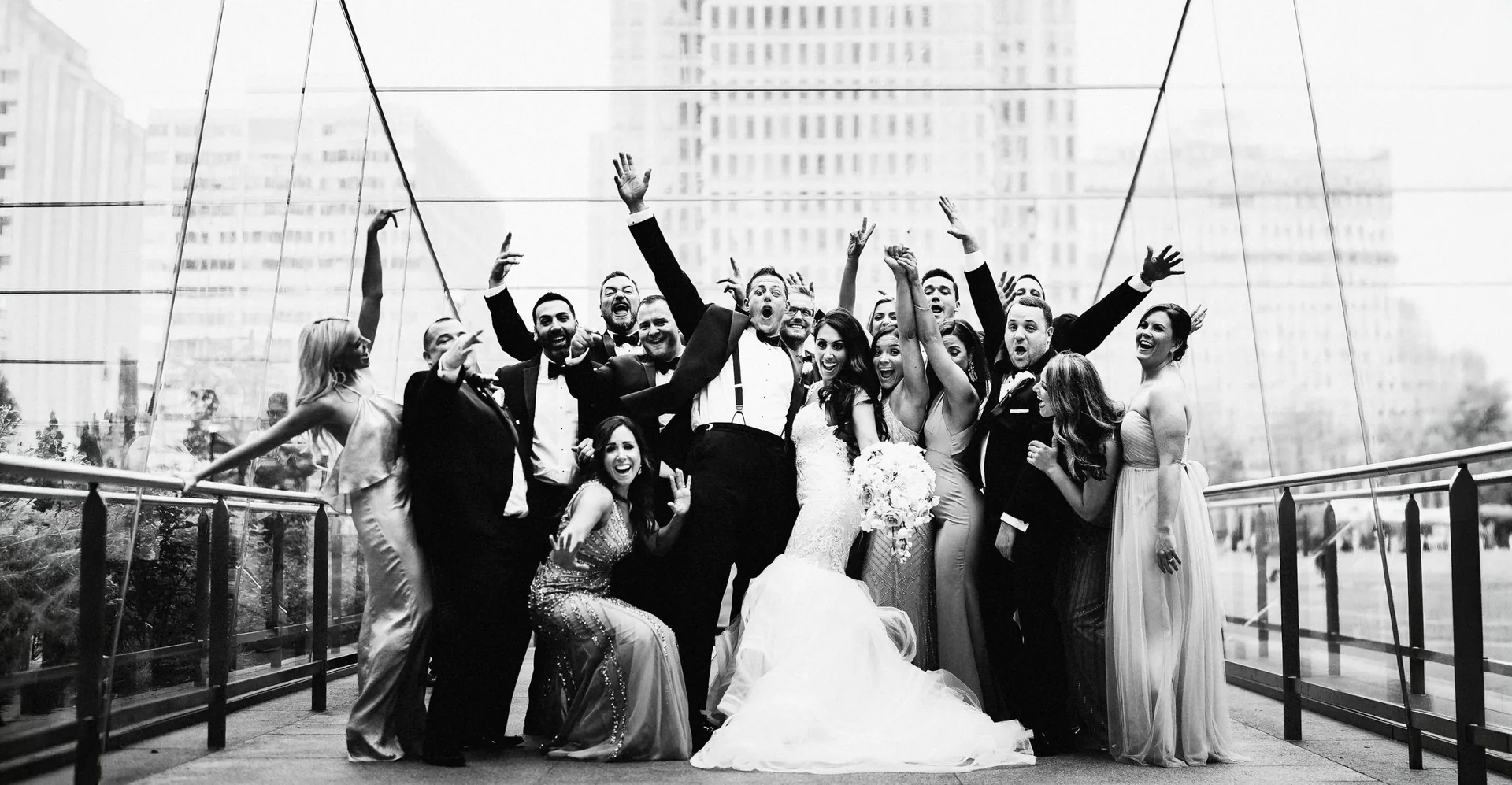 Black and white photo of a bride, groom and their wedding party cheering with arms raised on a glass city bridge
