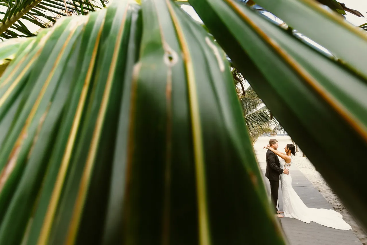 Bride and groom embracing on the beach boardwalk, framed by palm leaves, at ICONA Beach Resort in Wildwood, NJ