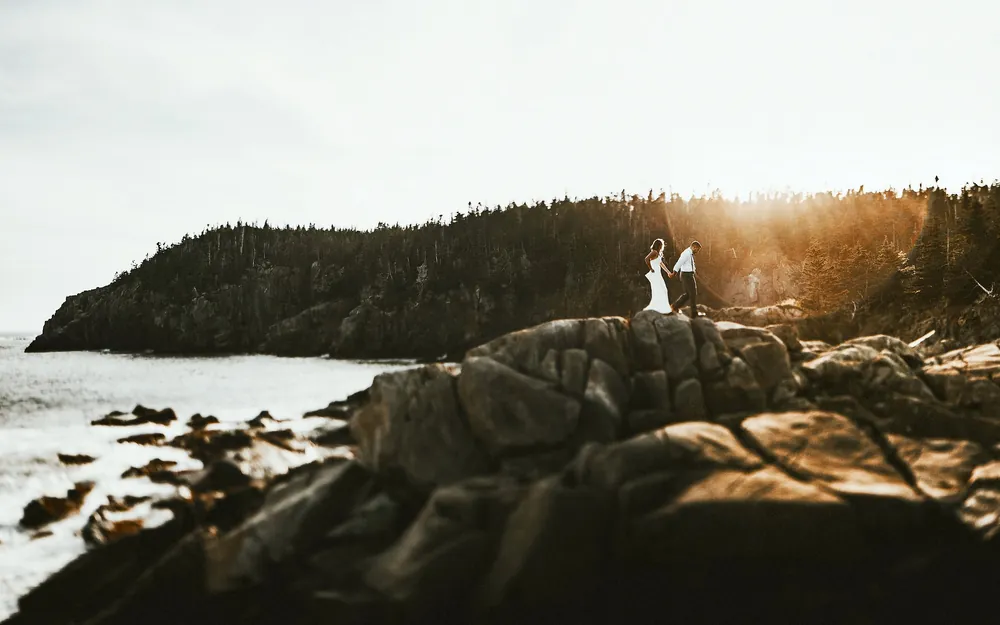 Bride and groom walking hand in hand across rocky sea cliffs on the coast of Maine at sunset