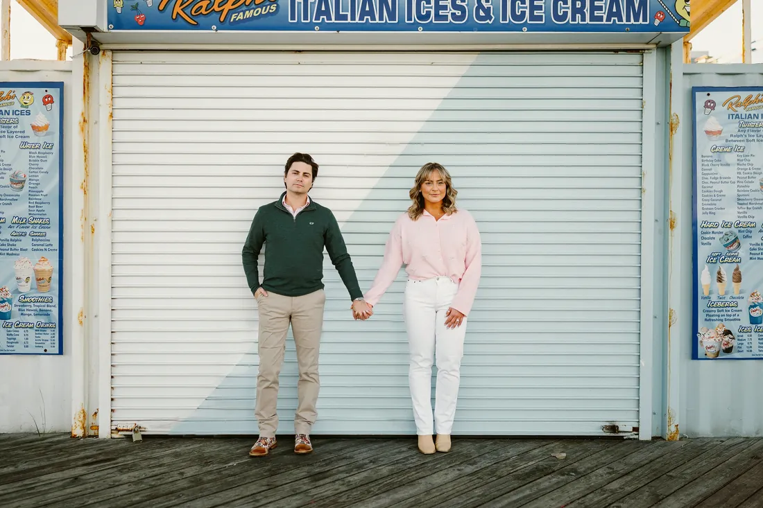 Engaged couple holding hands on the boardwalk in front of an Italian ice stand