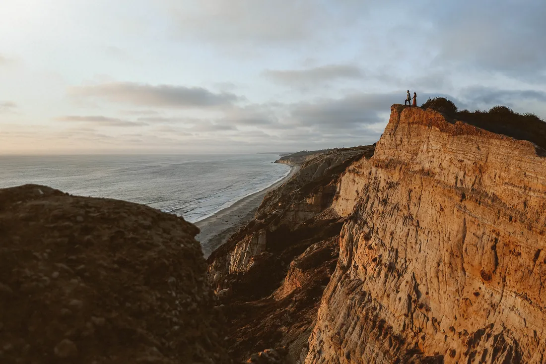 Engaged couple standing small on a cliff above the ocean at sunset