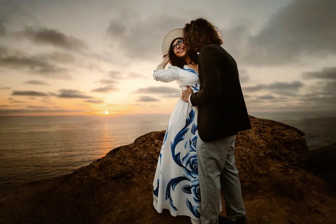 Engaged couple embracing on a cliff by the sea at sunset