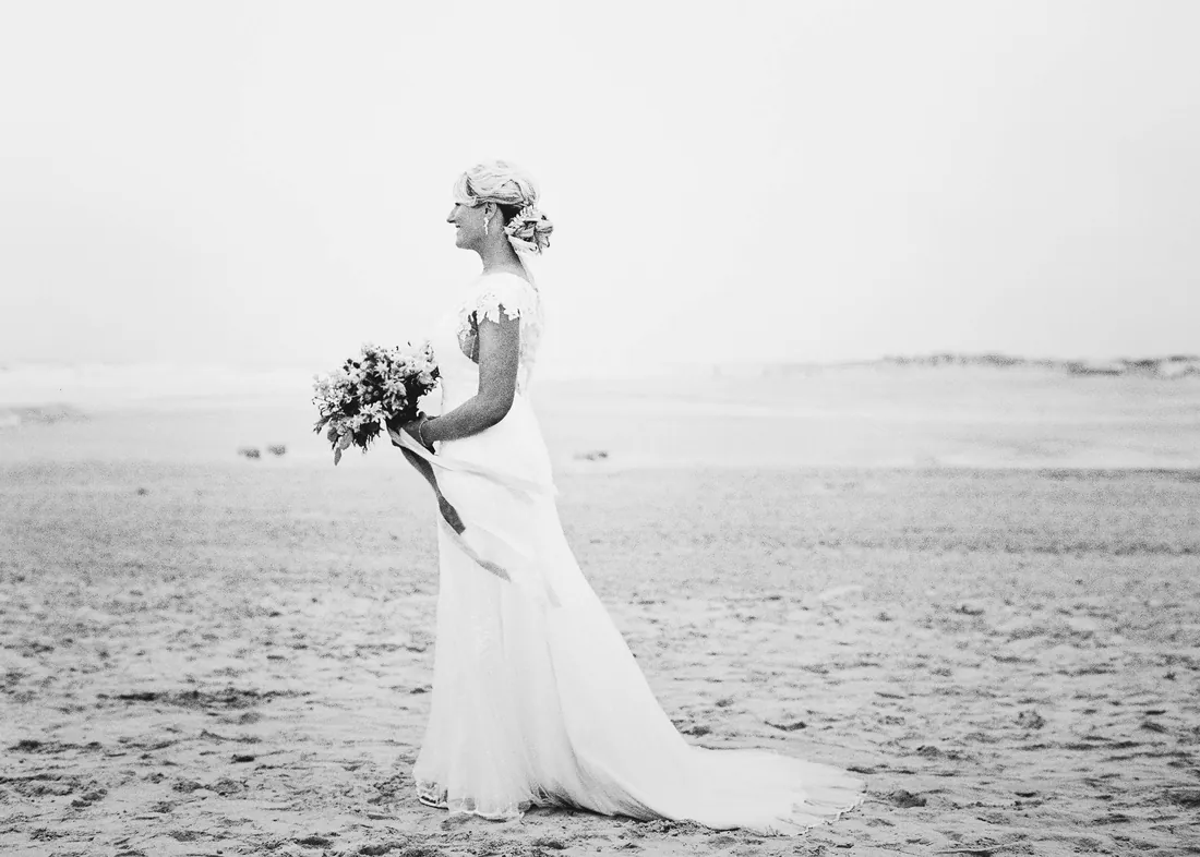 Black and white film photograph of a bride with her bouquet on the beach