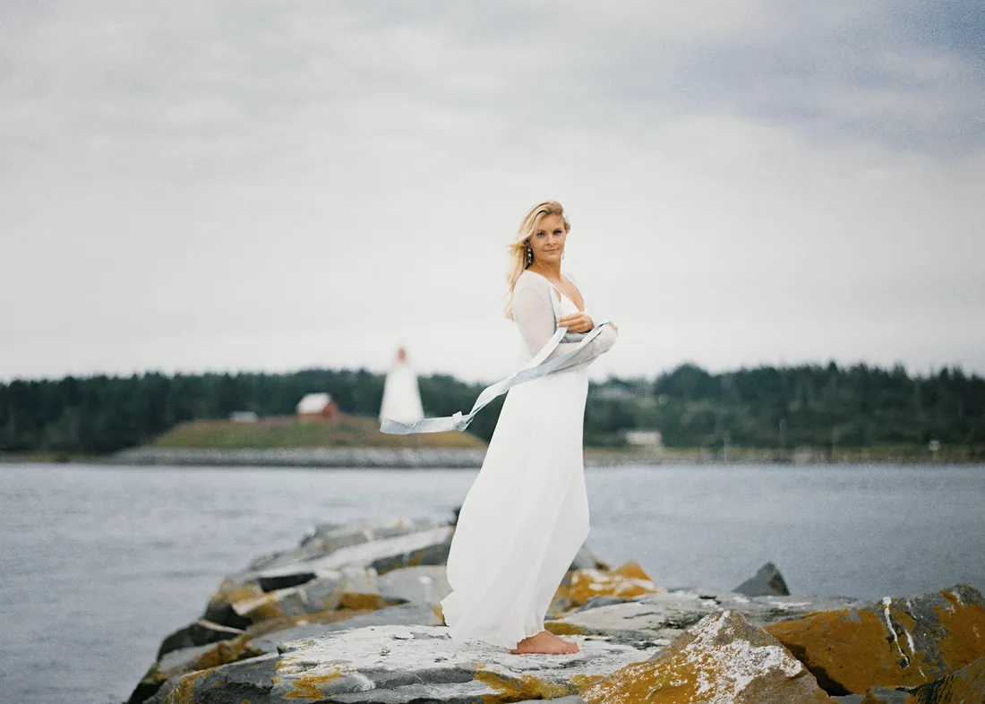 Film photograph of a bride standing on coastal rocks with a lighthouse in the distance