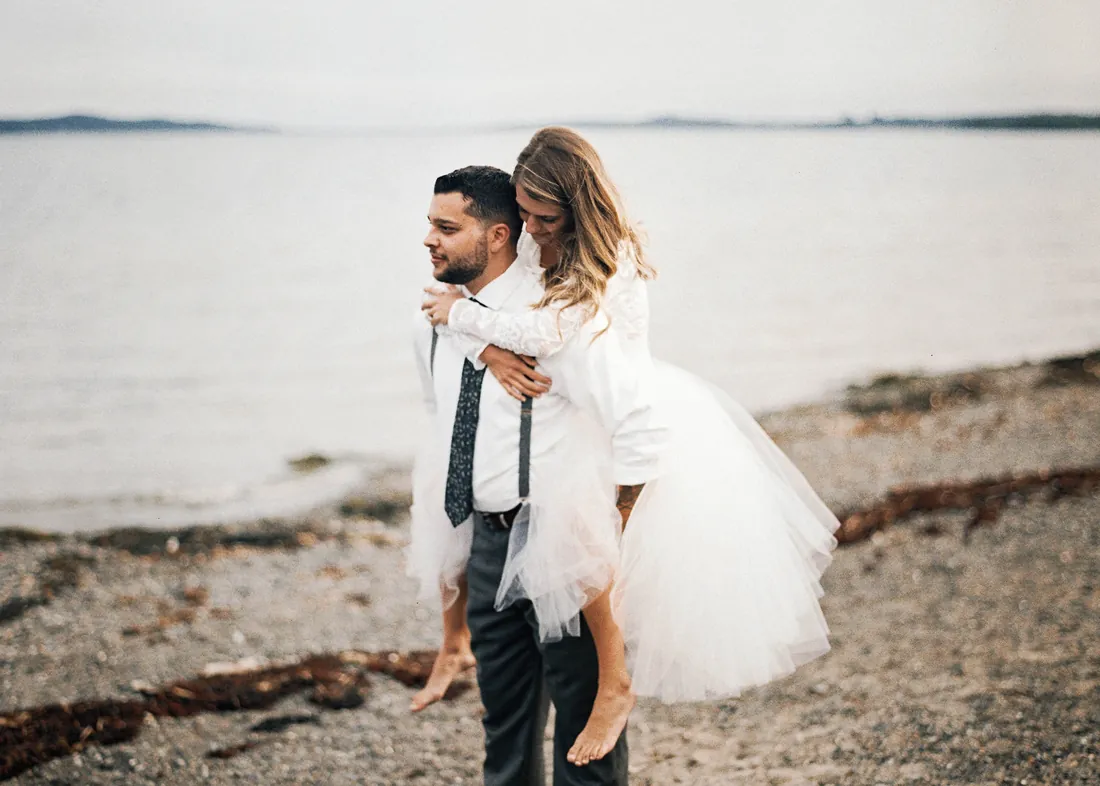 Film photograph of a groom giving his bride a piggyback ride on the beach