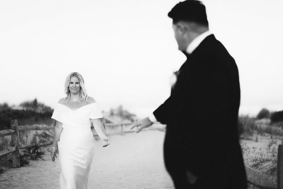 Black and white photo of a groom reaching for his bride on the beach