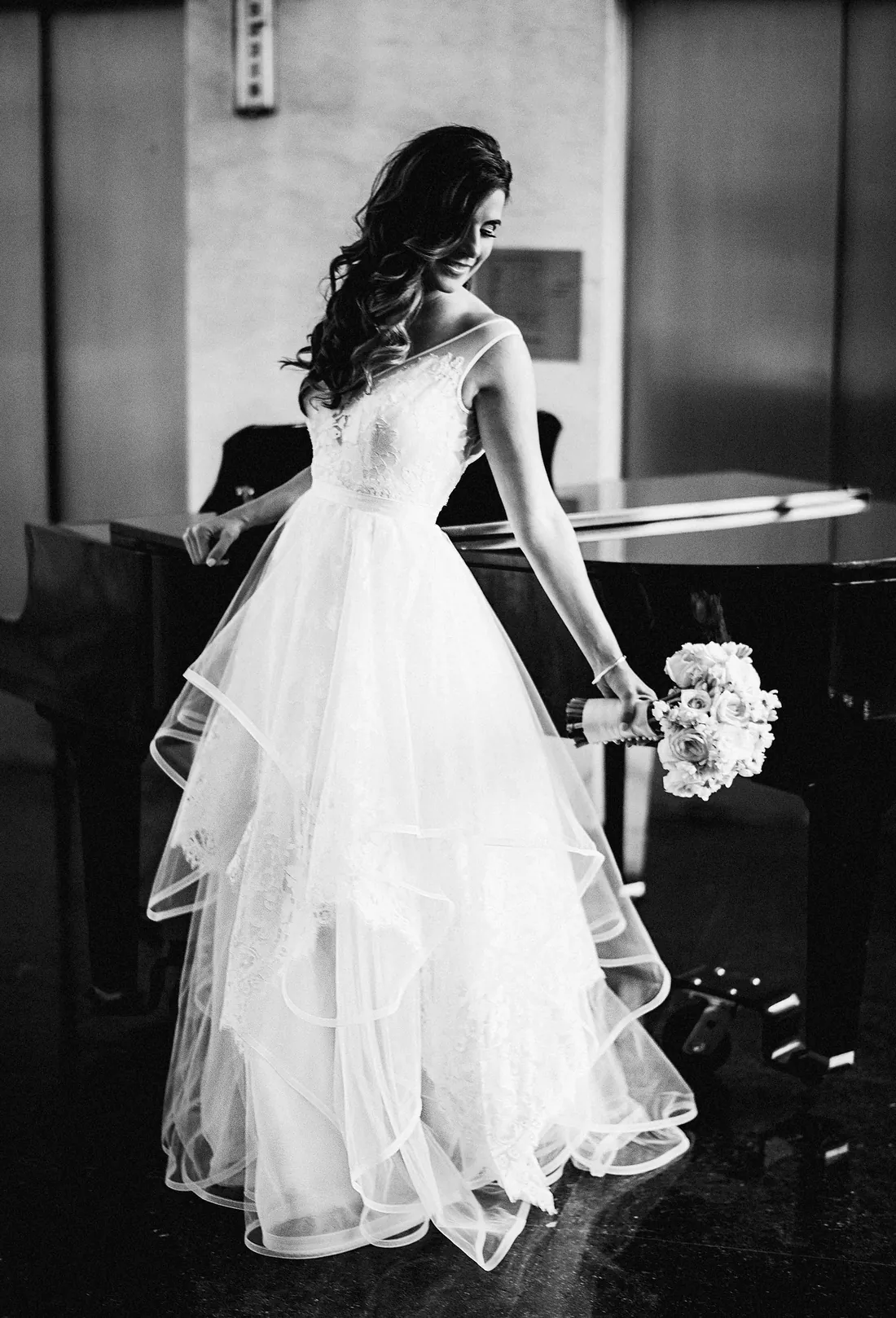 Black and white photo of a bride in a layered gown by a piano