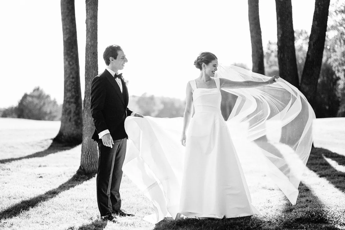 Black and white photo of a bride's veil blowing in the wind as her groom watches