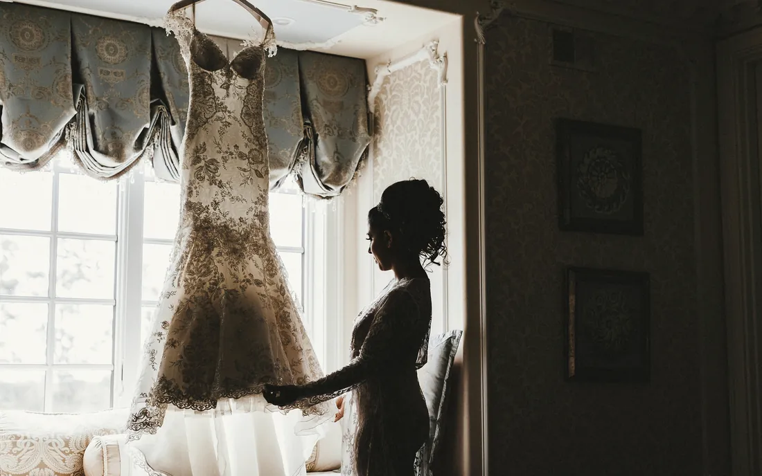 Bride in silhouette by a window with her gown hanging beside her