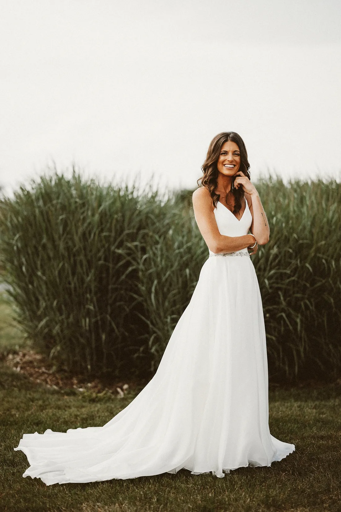 Smiling bride in a flowing gown in front of tall grass