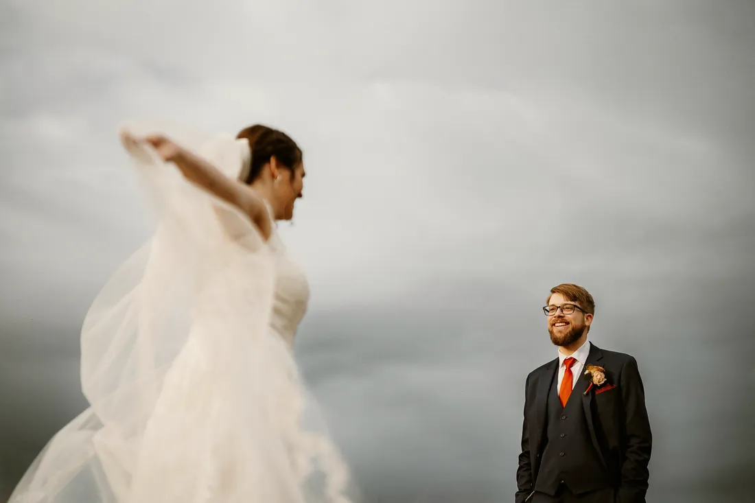 Bride twirling her gown under a gray sky as her groom watches