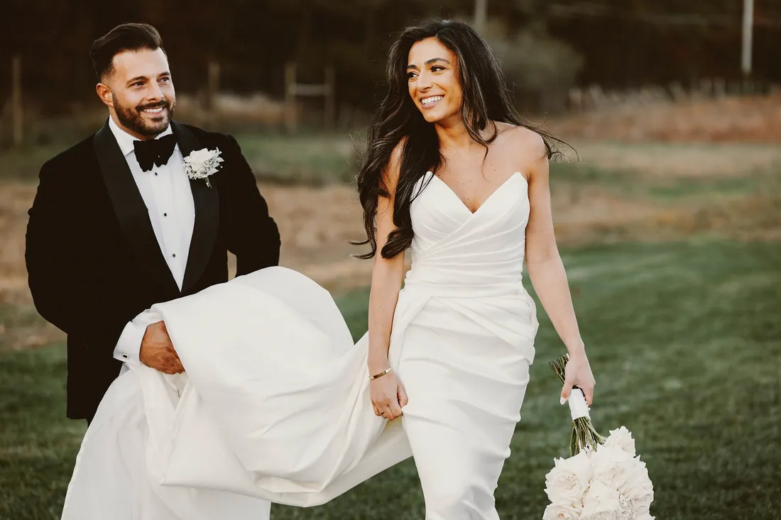 Groom carrying his bride's train as they walk through the grass