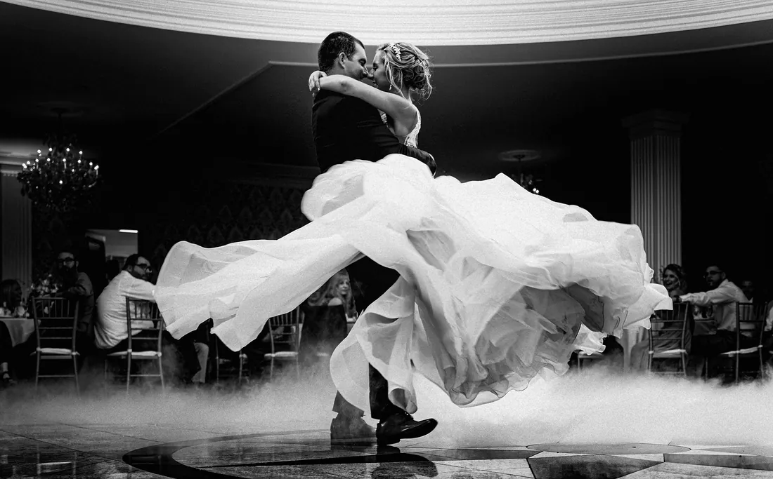 Black and white photo of a groom lifting his bride on a fog-covered dance floor