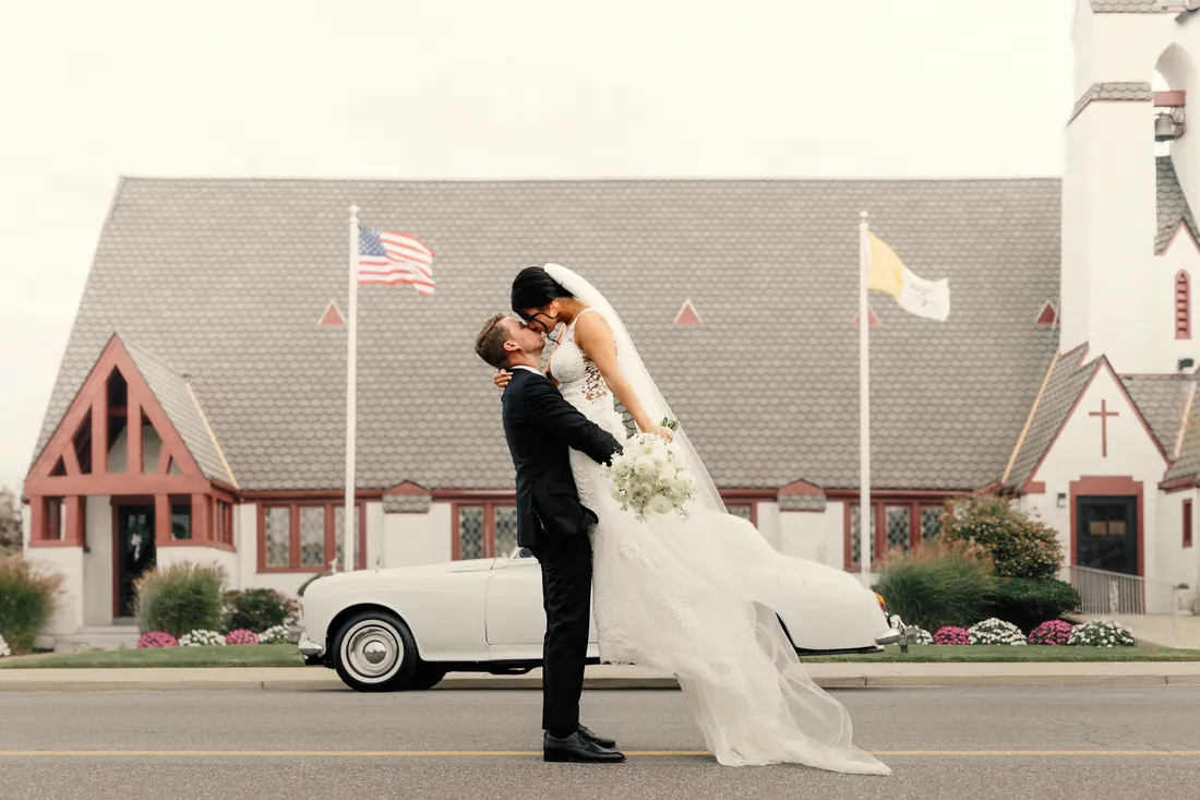 Groom lifting his bride for a kiss outside the church beside a vintage car