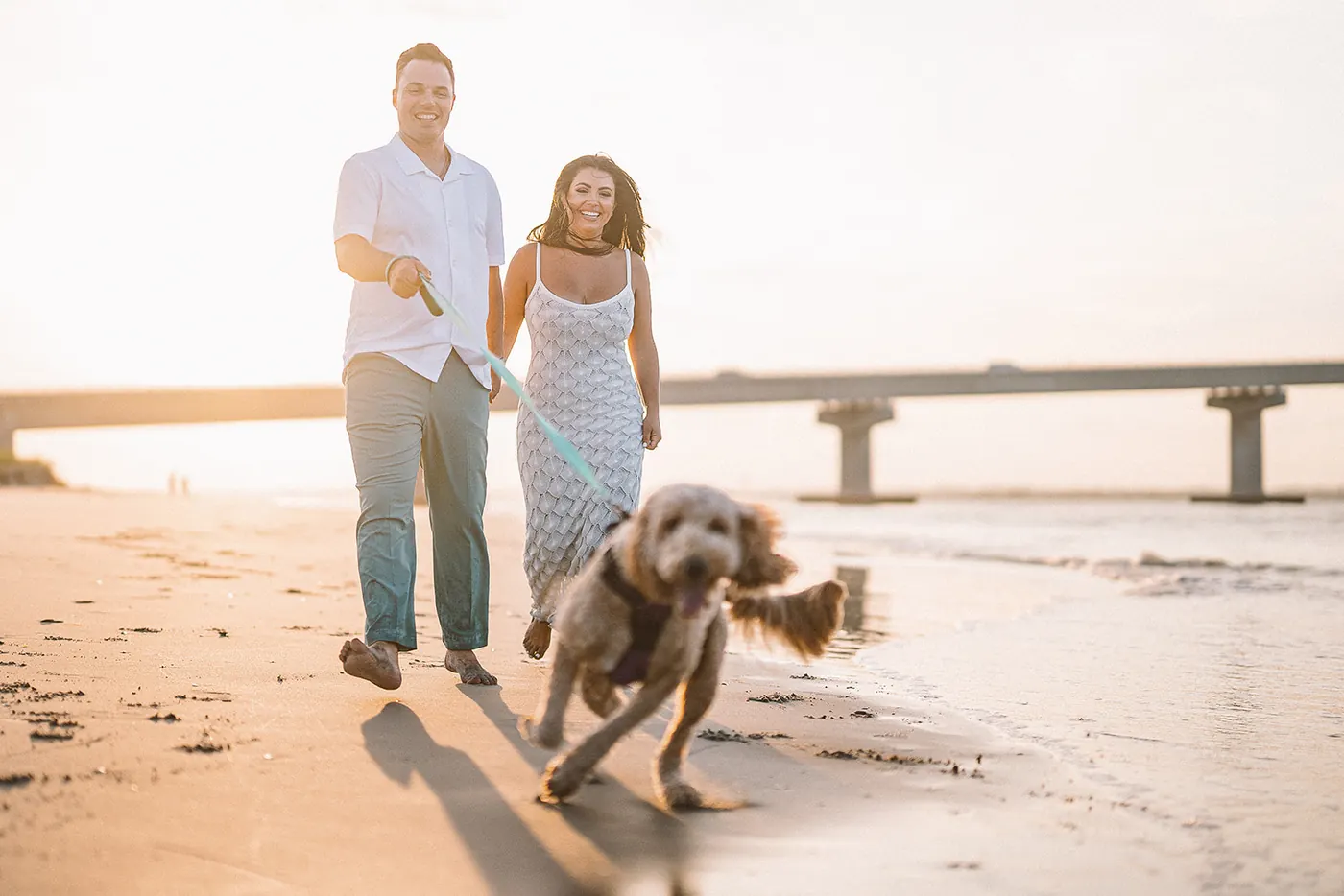Branon and Celeste walking their goldendoodle along the beach at golden hour in Ocean City, NJ, near the Longport bridge