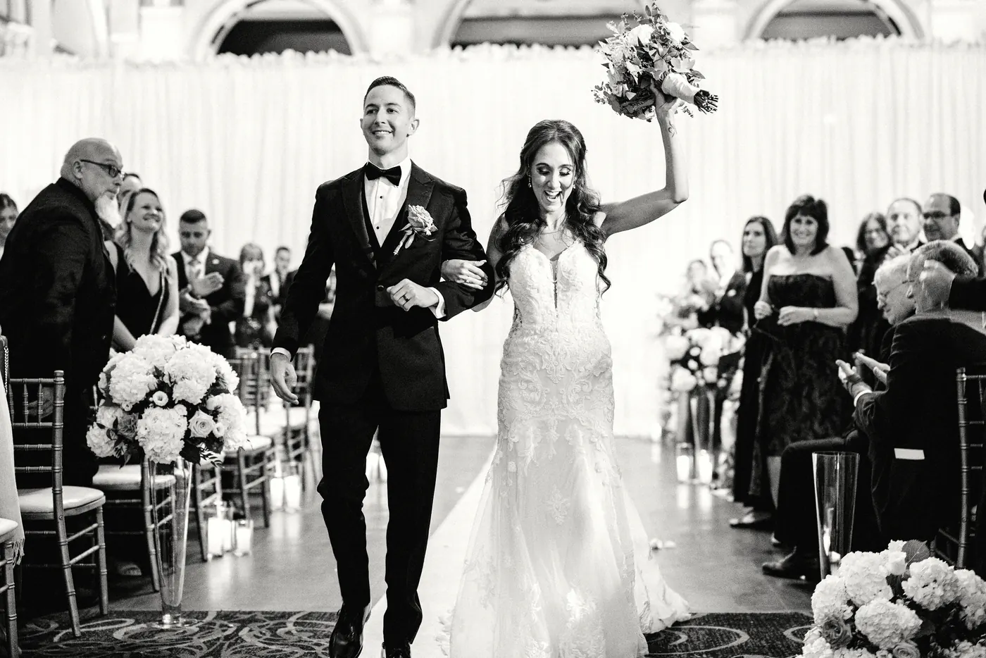 Black and white photo of Danielle raising her bouquet as they walk back up the aisle at Cescaphe Ballroom at the Ben in Philadelphia
