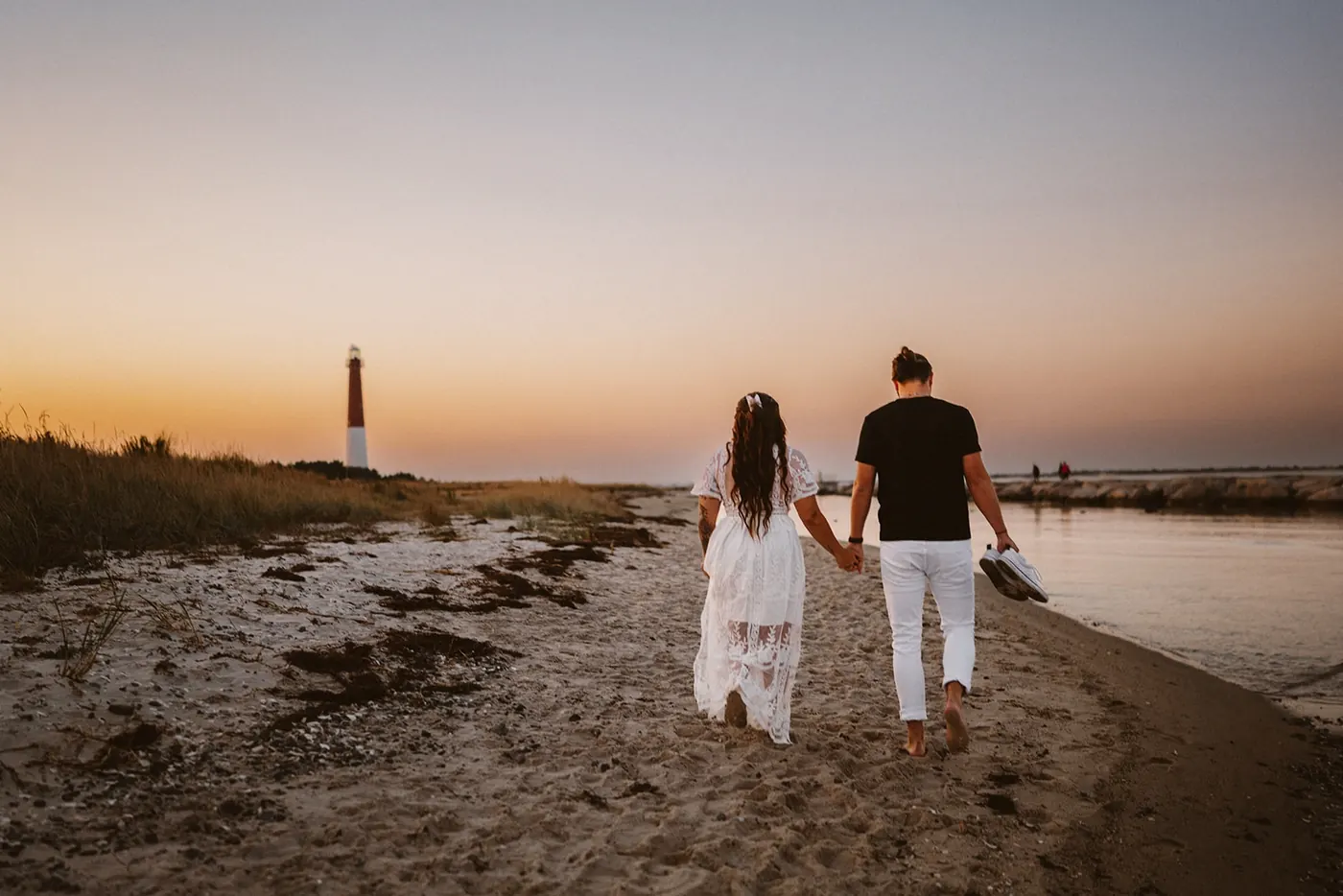 Dan and Madison walking hand in hand along the bay beach toward Barnegat Lighthouse at dusk