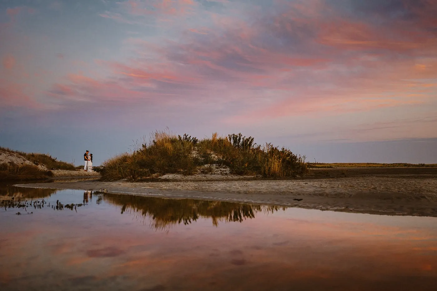 Dan and Madison small in the landscape beside a dune and a pink sky reflection near Barnegat Lighthouse on Long Beach Island, NJ