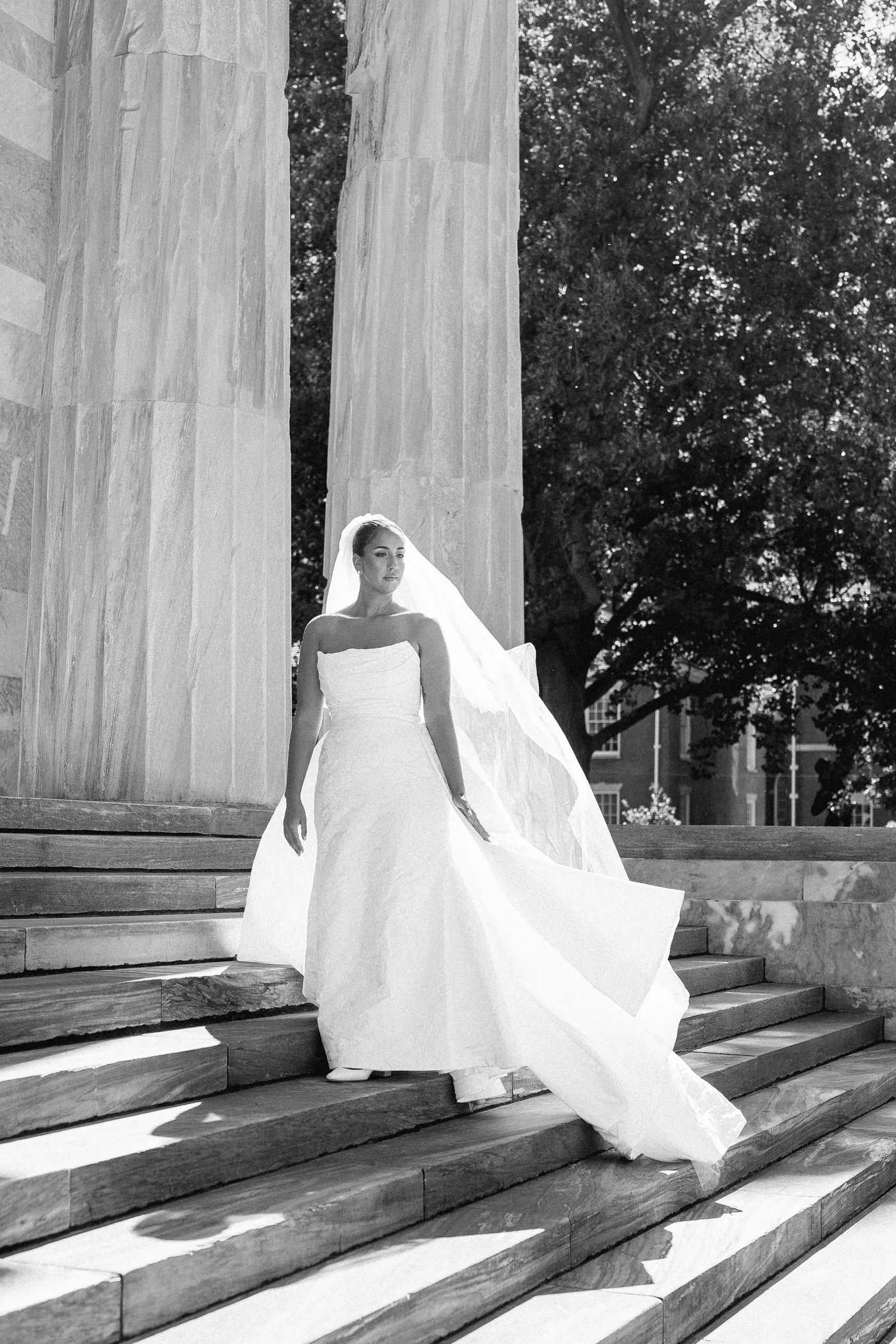 Black and white photo of Gianna walking down marble steps between tall columns, her veil trailing
