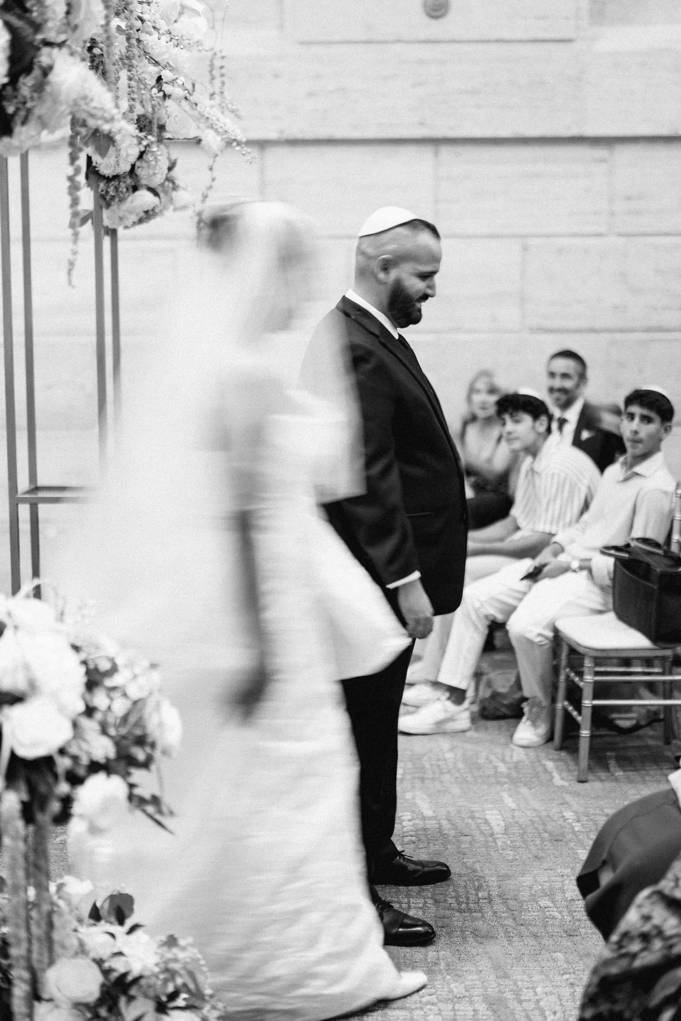 Black and white photo of Gianna circling Simon under the chuppah as guests smile