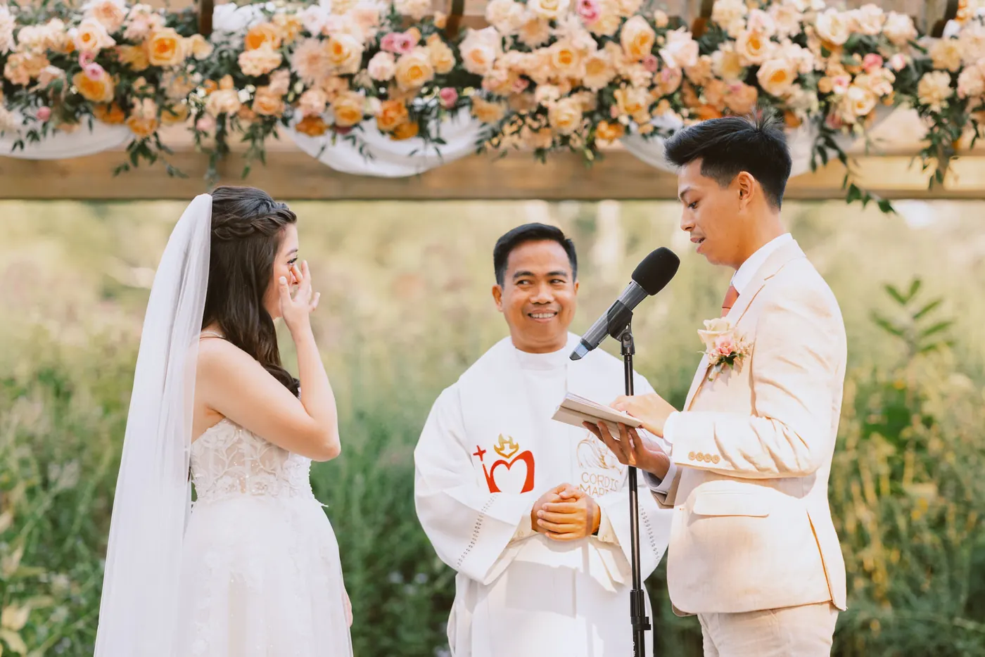 Ivan reading his vows as Camille wipes away a tear under the peach-flowered pergola