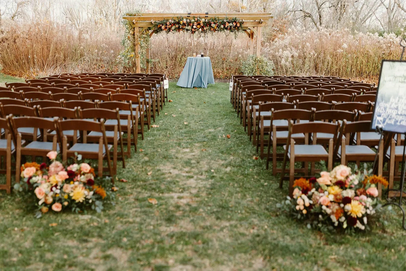 Rows of wooden chairs and autumn flowers lining the aisle to the floral pergola for Jimmy and Kelsey's ceremony at Bishop Farmstead