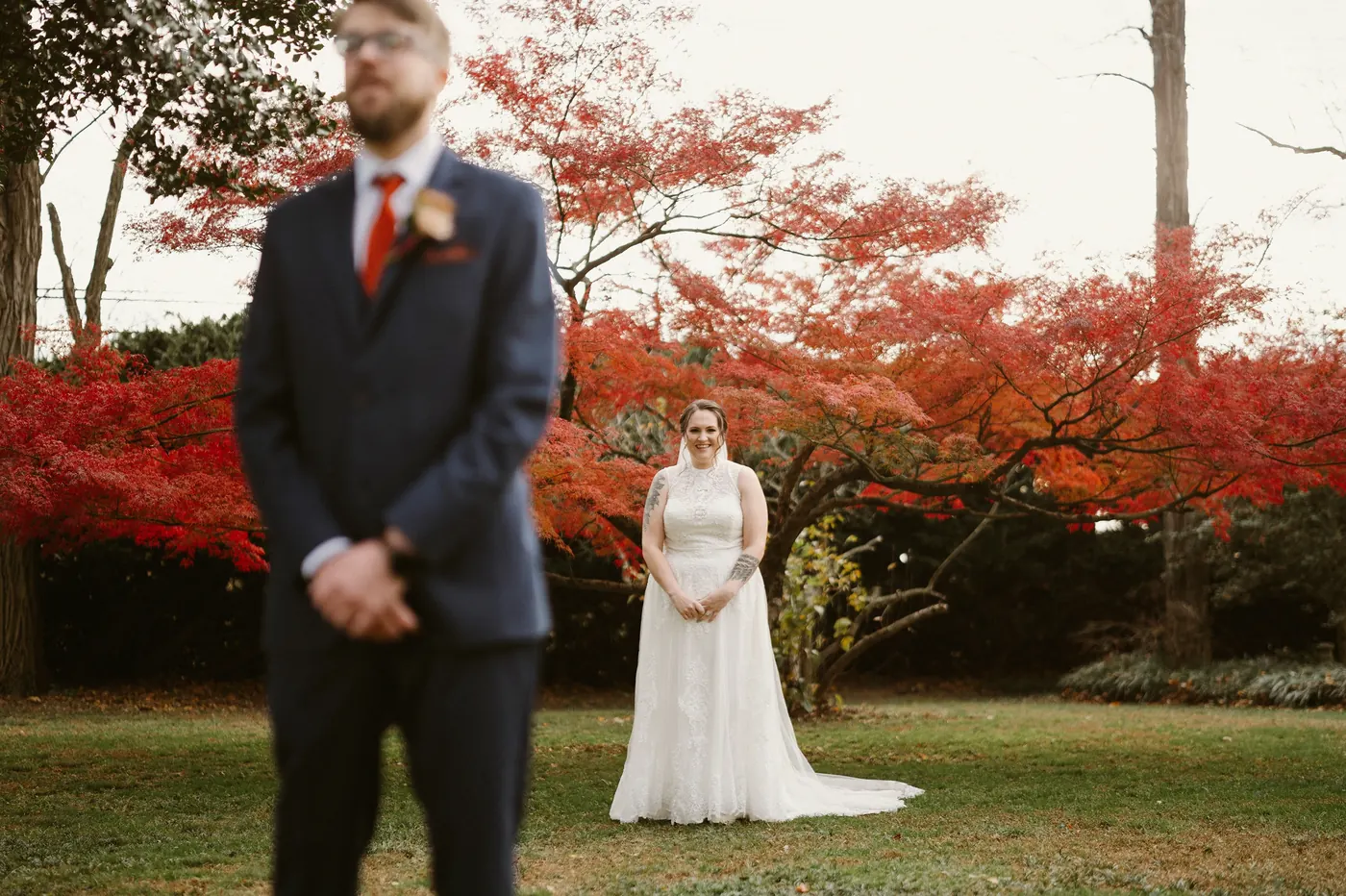 Kelsey walking up behind Jimmy for their first look in front of a red Japanese maple at Bishop Farmstead