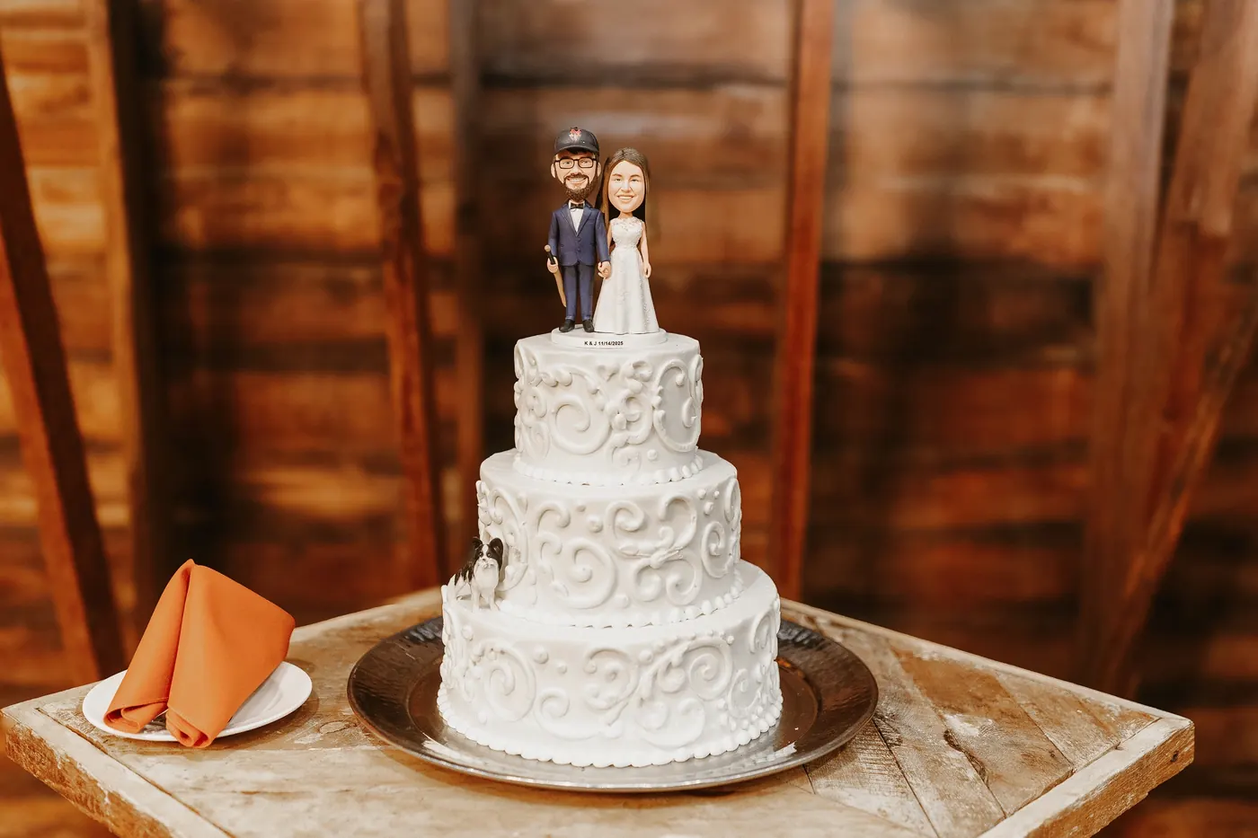 Three-tier white wedding cake with a custom bobblehead topper in the barn