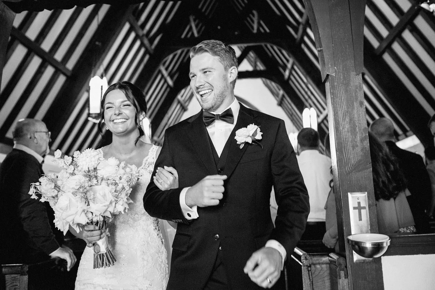 Black and white photo of Mason and Morgan beaming as they walk up the aisle beneath the church's wooden beams