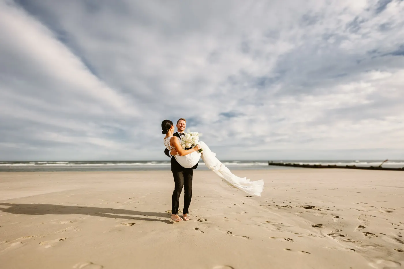 Mason carrying Morgan on the beach as her veil flies in the wind