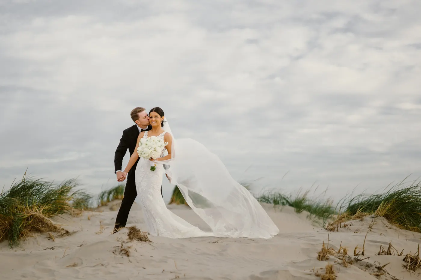 Mason kissing Morgan's cheek in the dune grass as her veil blows behind her