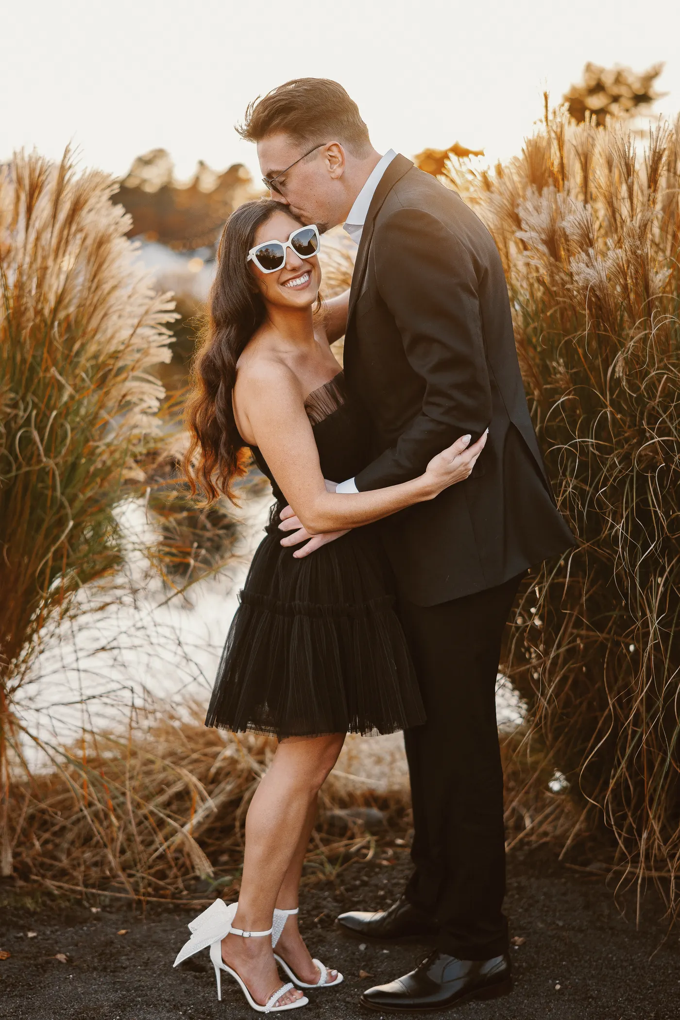 Morgan in a black dress and white heels hugging Will beside tall ornamental grass at Renault Winery in Egg Harbor City, NJ