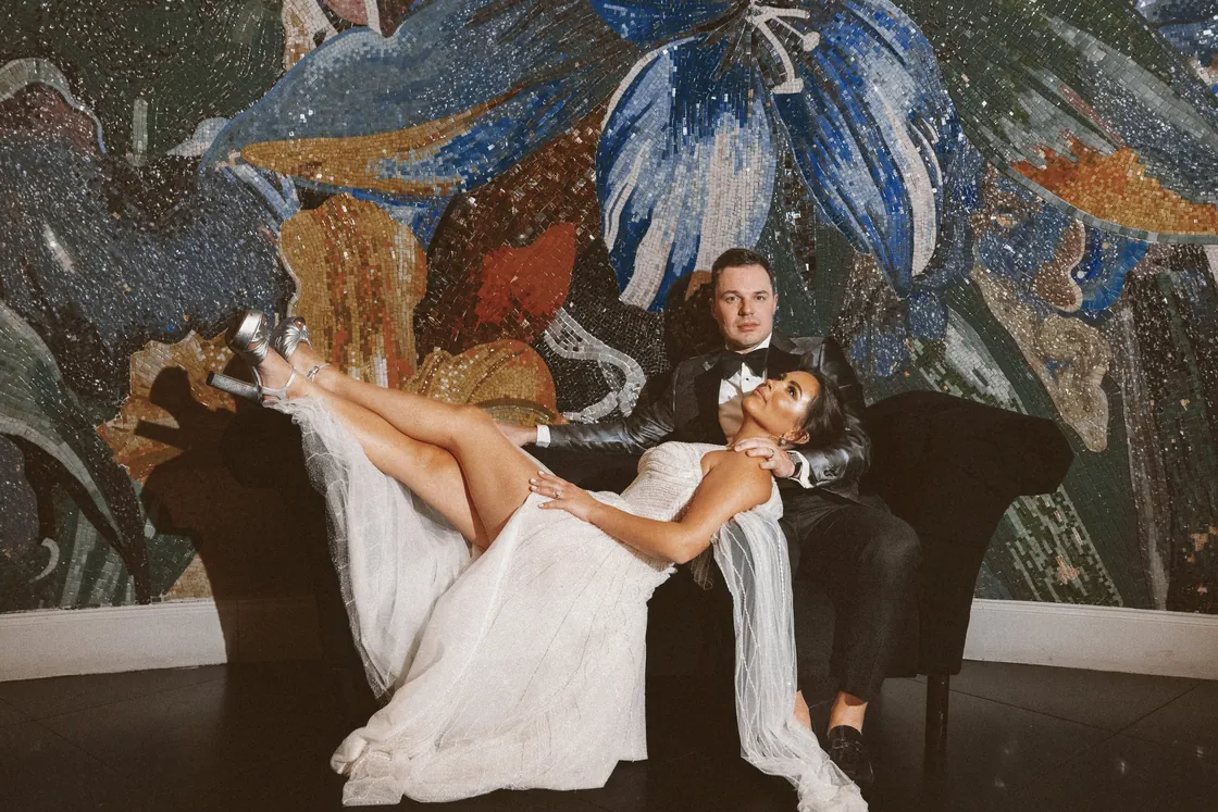 Branon and Celeste posing on a black velvet chaise in front of a colorful floral mosaic at The Vie by Cescaphe in Philadelphia